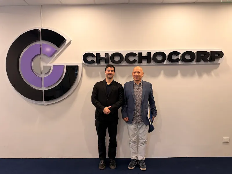 Dois integrantes da Chochocorp junto ao logo na recepção